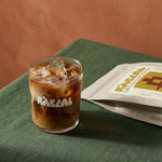 iced coffee in rascal branded glass with terracotta background and green linen table cloth and bag of rascal copy next to it