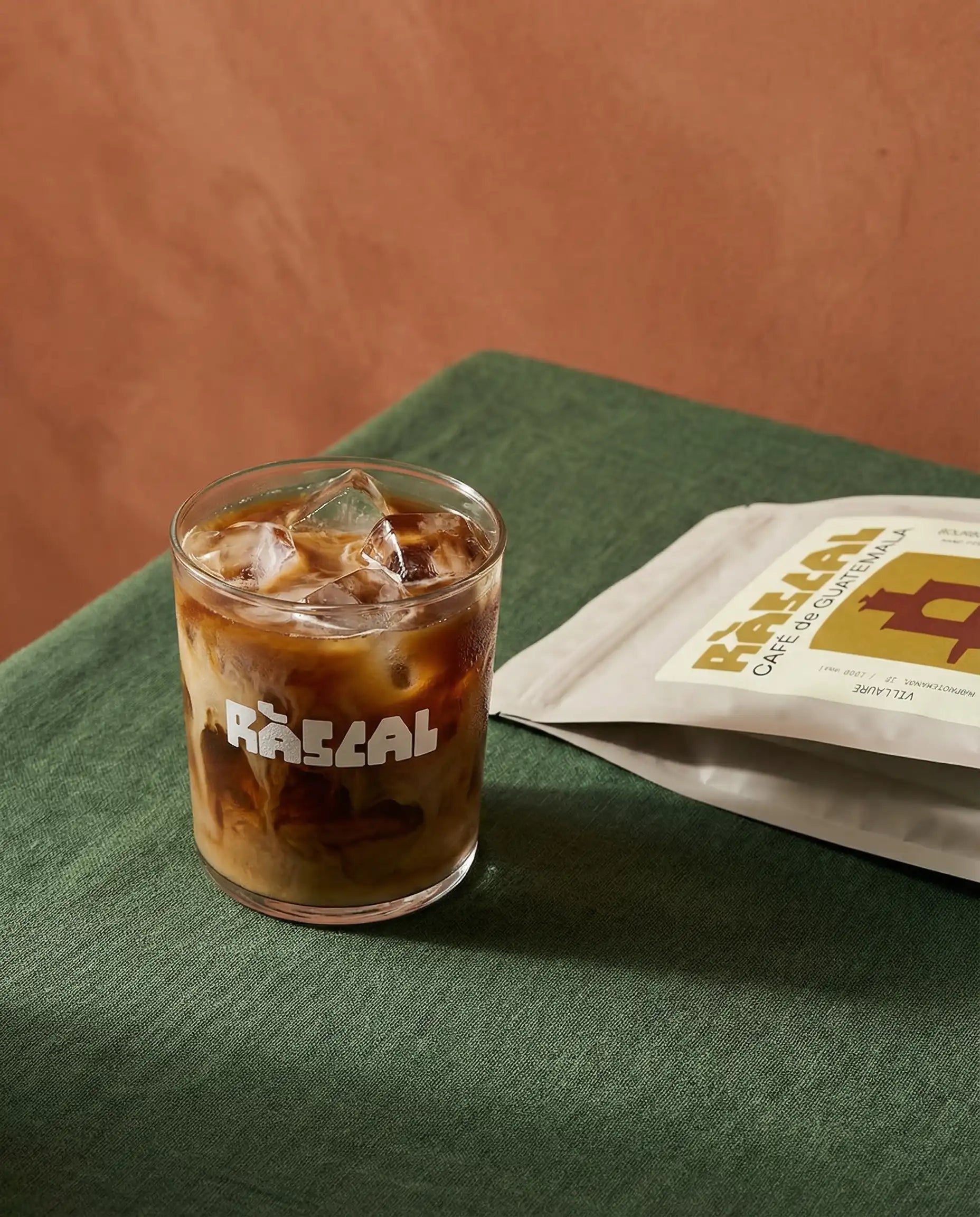 iced coffee in rascal branded glass with terracotta background and green linen table cloth and bag of rascal copy next to it